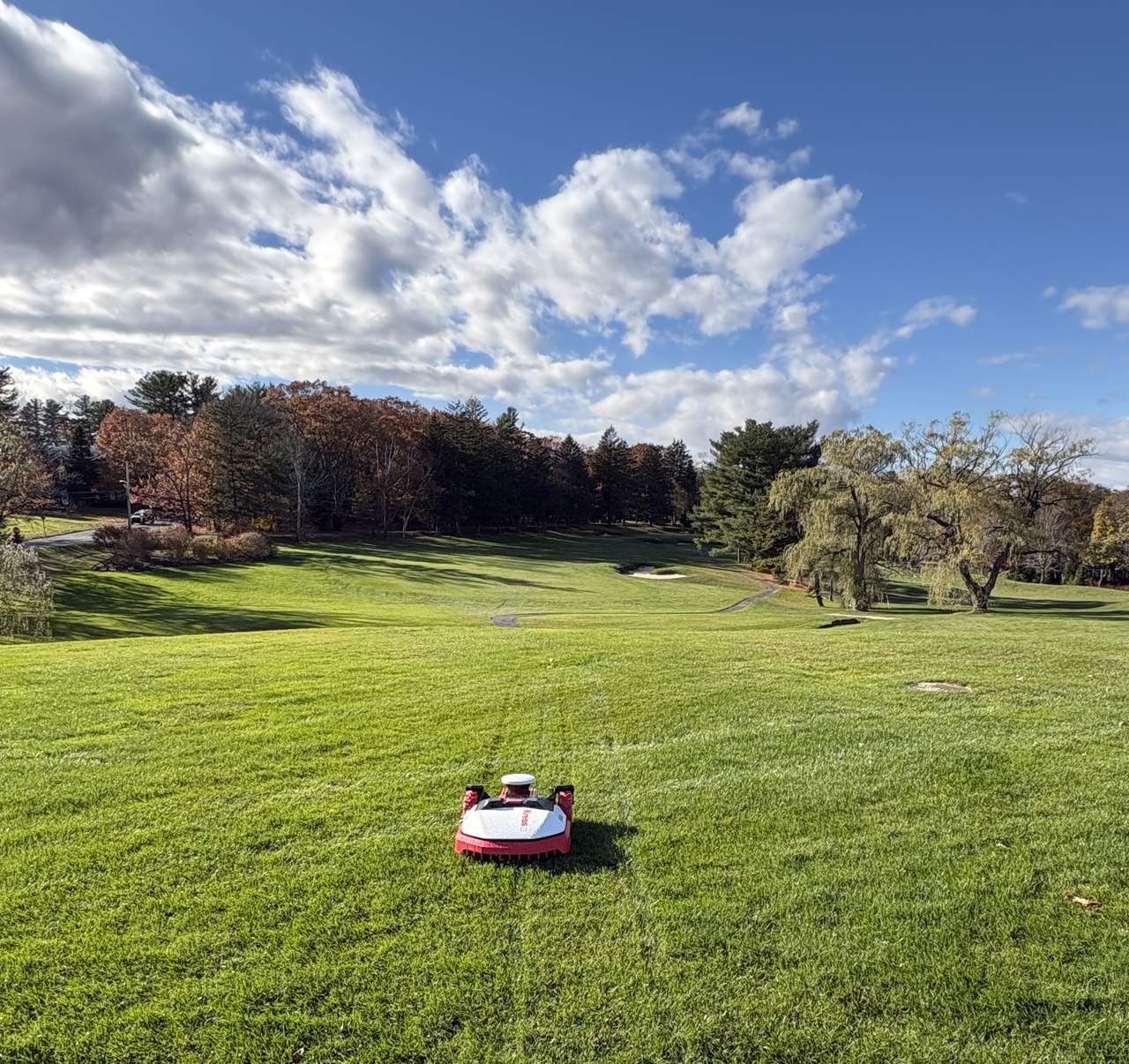 A red Kress robot mowing a fairway in fall, with orange foliage and a big blue sky